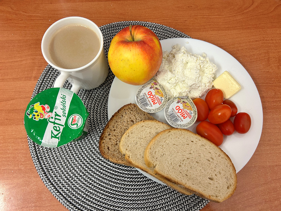 Na zdjęciu znajduje się: Kefir 2% tł., Kawa zbożowa z mlekiem/p, Chleb mieszany pszenno-żytni, Chleb Graham, Masło extra 82%, Miód, Twarożek, Jabłko, Pomidorki koktajlowe